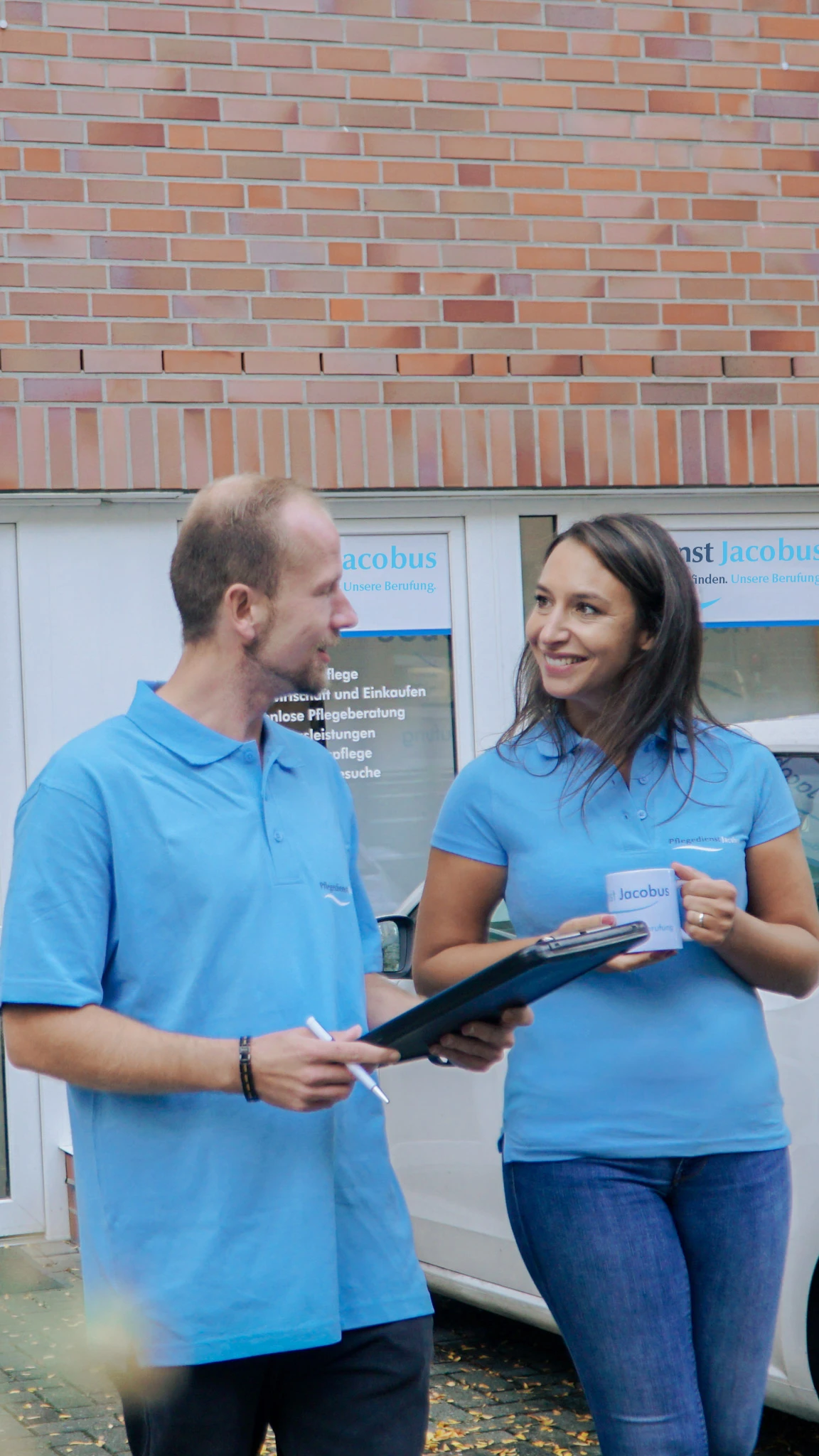 Zwei Pflegekräfte in blauen Poloshirts unterhalten sich vor einem Gebäude, eine hält eine Tasse, der andere ein digitales Tablet.