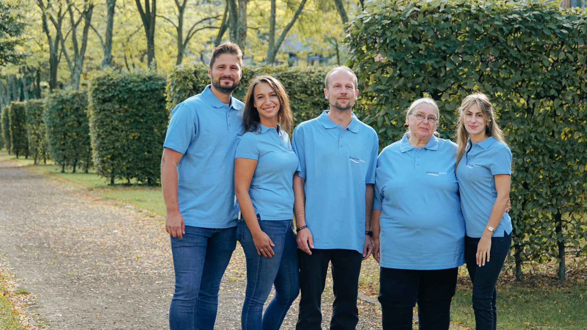 Fünf Mitarbeiter von Pflegedienst Jacobus stehen im Park, tragen hellblaue Poloshirts und lächeln in die Kamera.