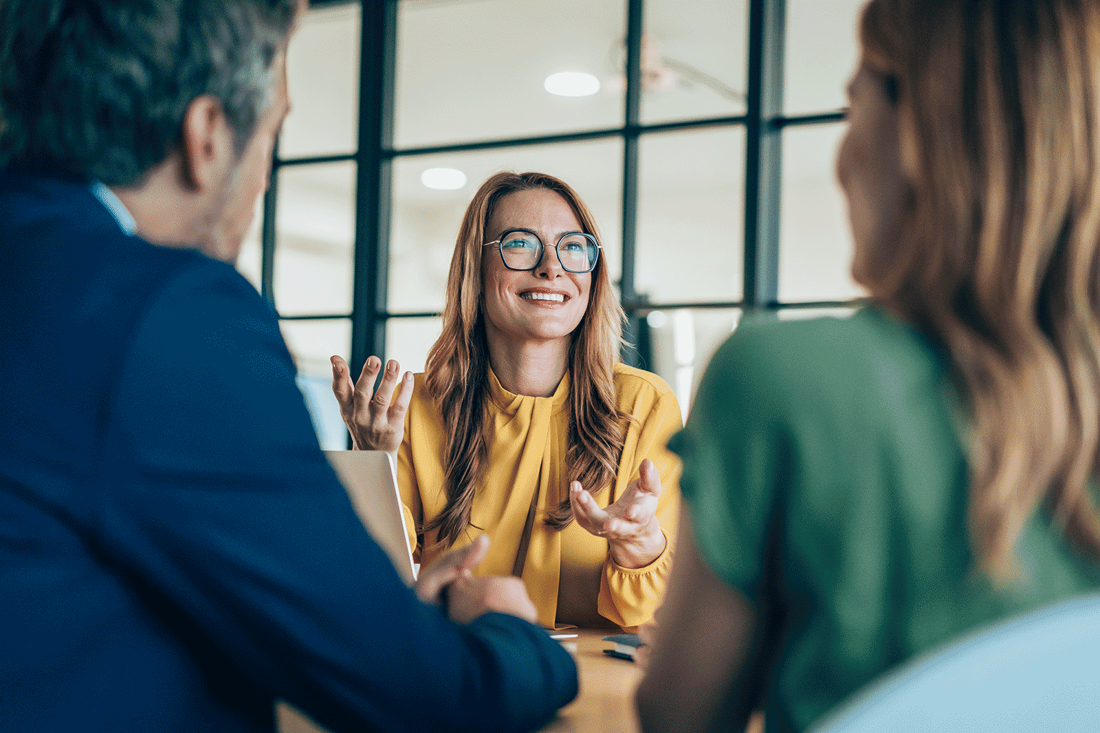 A woman in a yellow blouse and glasses smiling and gesturing while talking to two people in a modern office.