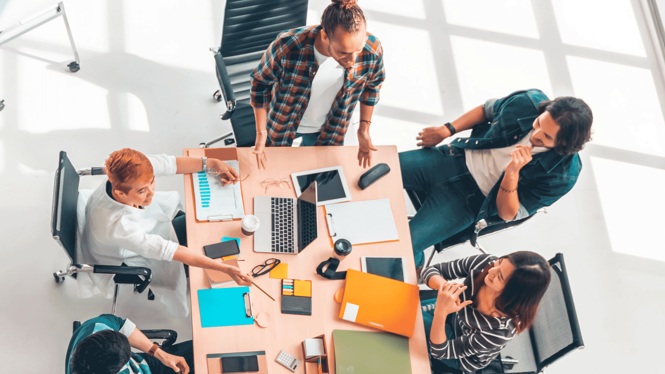 Top view of a diverse group of five people having a meeting around a table with laptops, tablets, and office supplies.