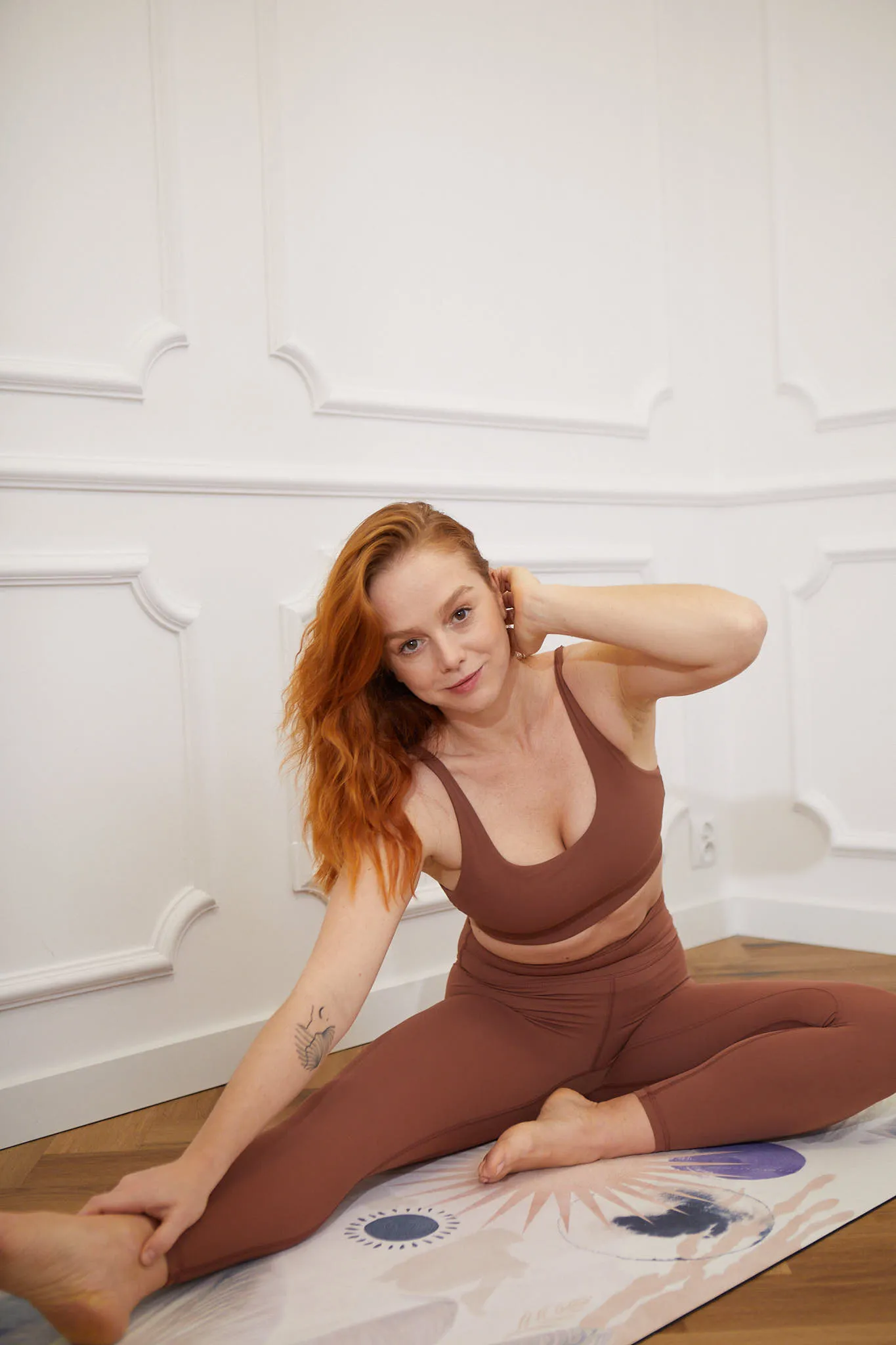 Red-haired woman in brown workout clothes seated on a pilates mat performing a seated hamstring stretch.