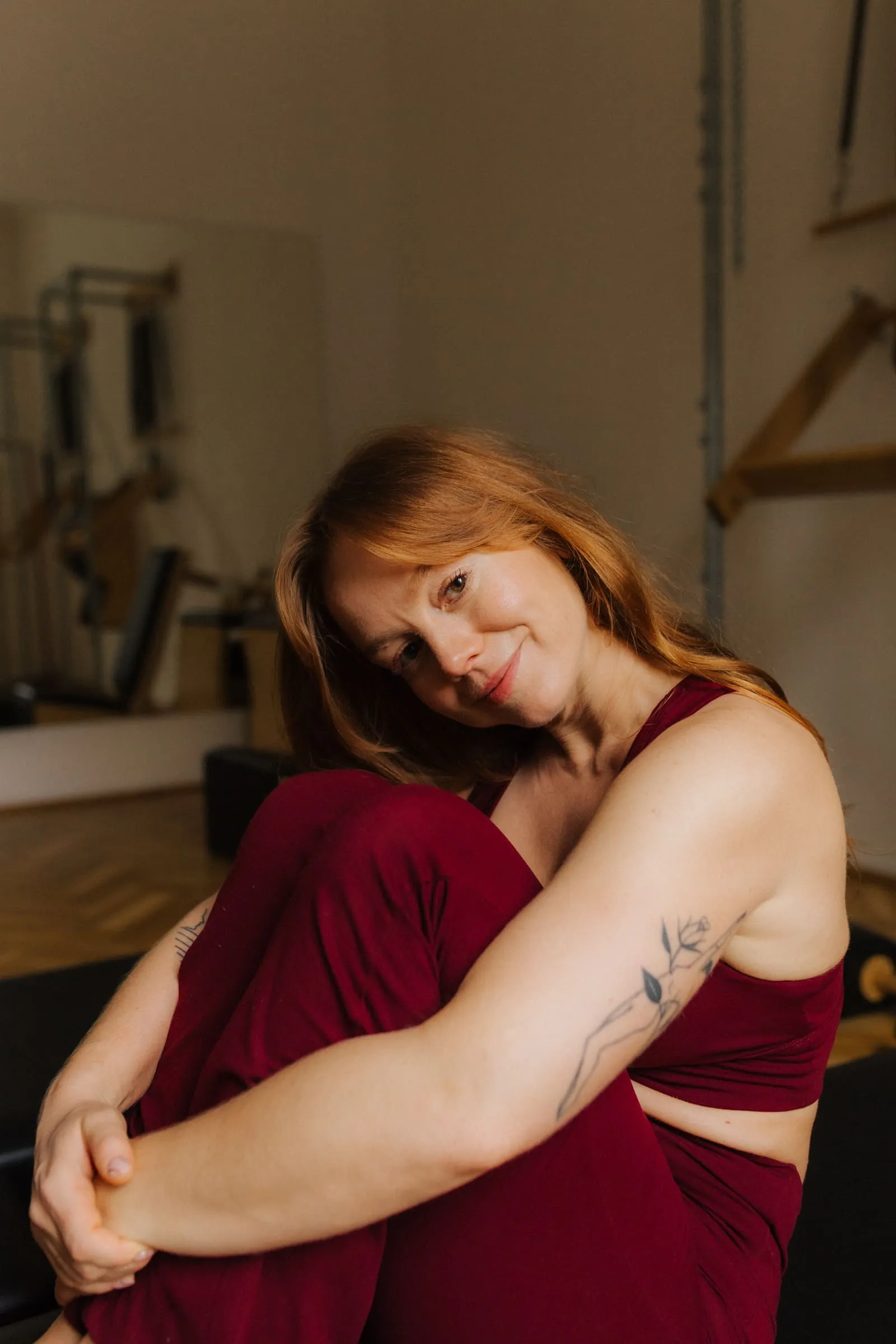 Smiling woman with red hair wearing a burgundy sports outfit sitting and hugging her knees indoors.
