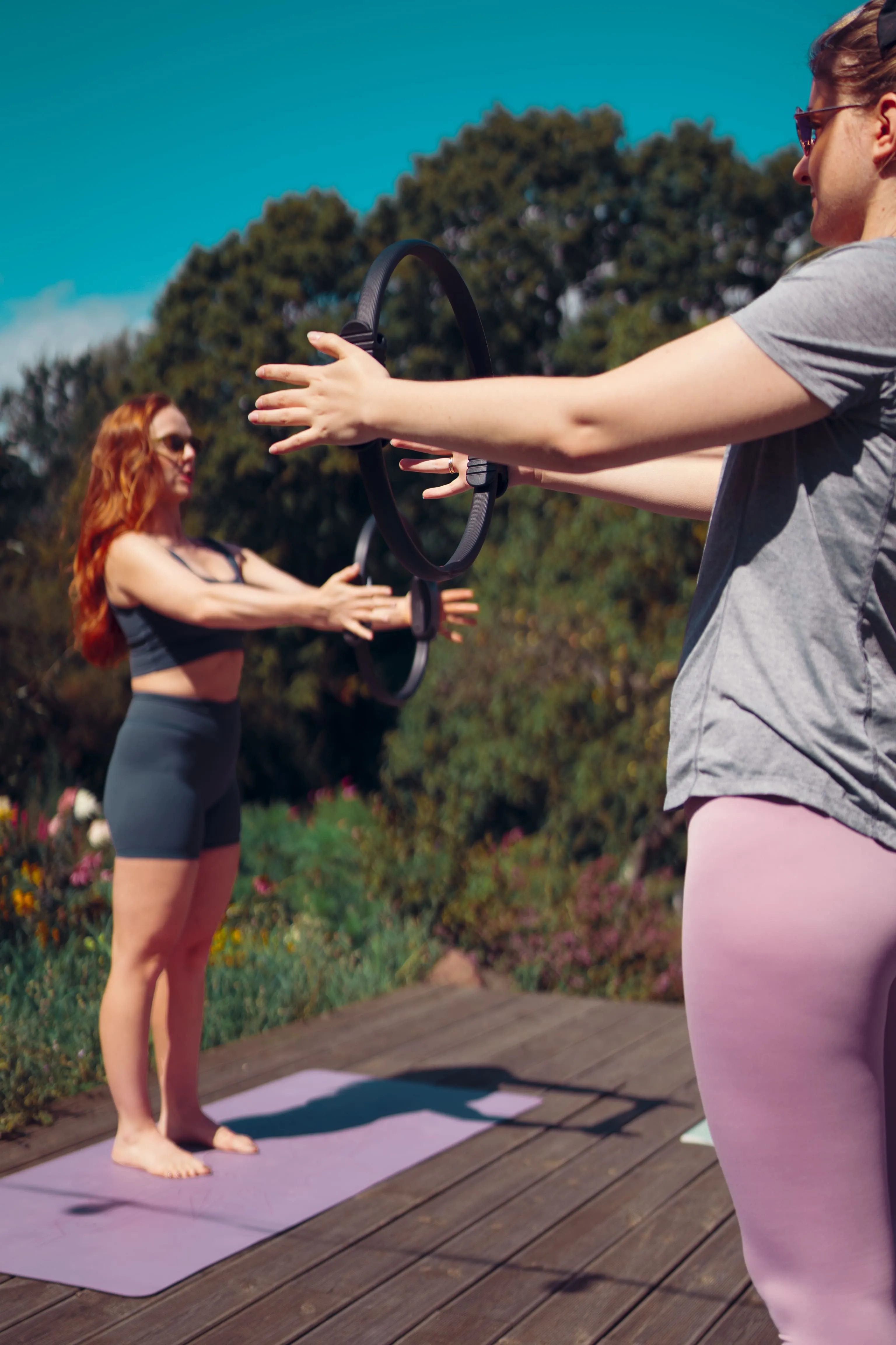 Two women exercising outdoors with Pilates rings on a wooden deck surrounded by greenery.
