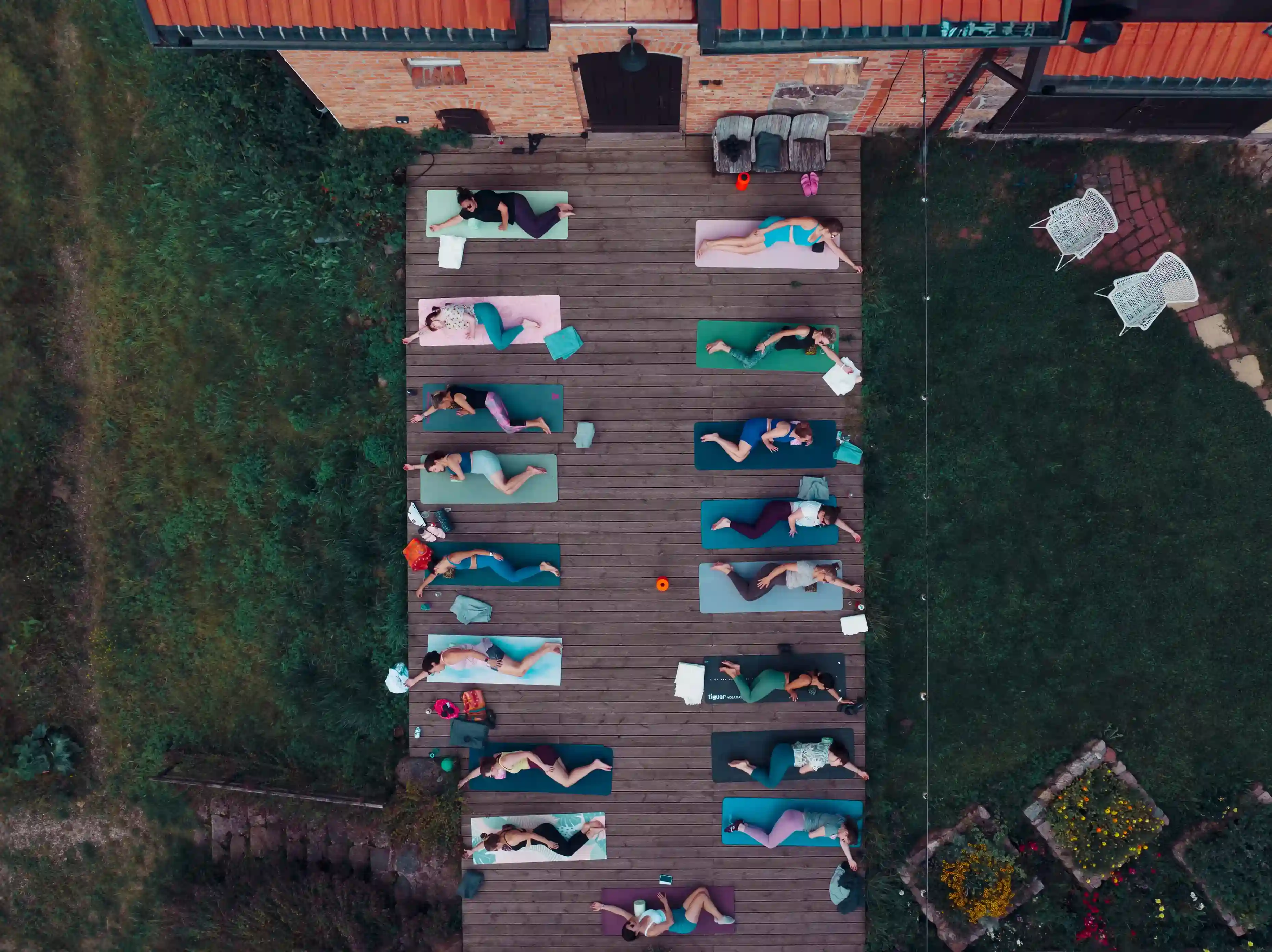Aerial view of people lying on yoga mats arranged in rows on a wooden deck outside a brick building, surrounded by grass and garden beds.