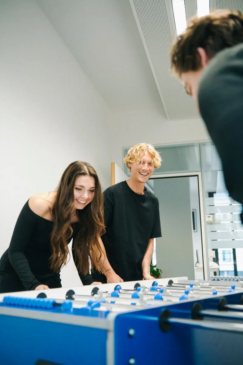 3 Personen stehend lachend an einem Kickertisch