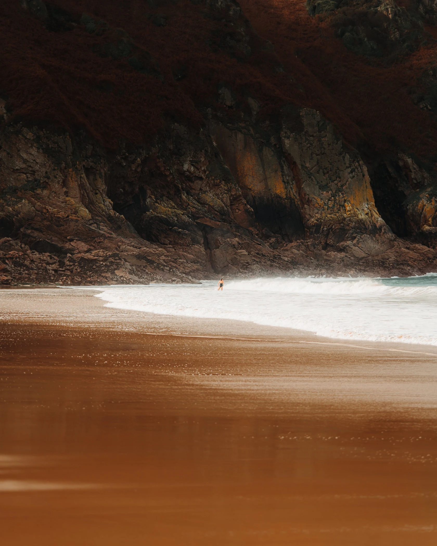 A solitary figure stands on a sandy beach with waves breaking near them, framed by a dramatic backdrop of a rugged cliff with reddish hues.