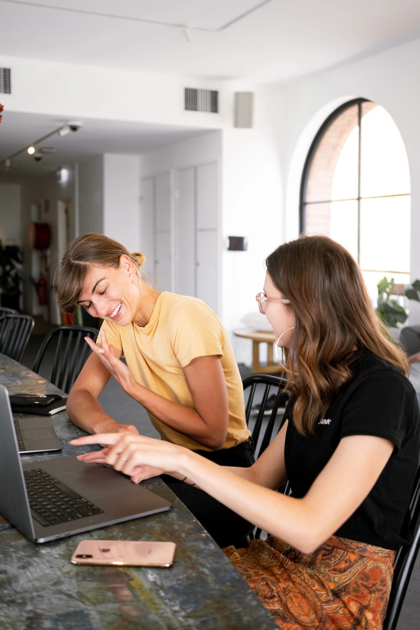 Two women engaged in a cheerful conversation at a table with a laptop, one gesturing animatedly, possibly in a discussion or collaborative work environment.