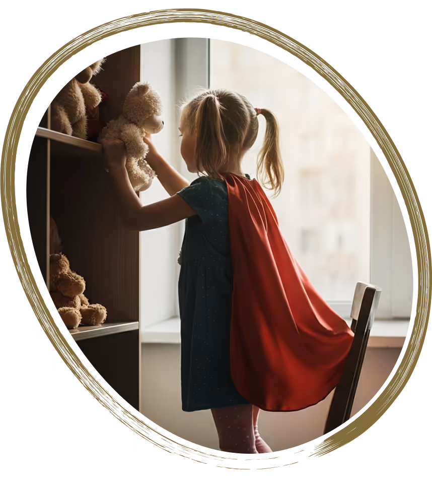 Girl wearing a red cape playing superhero, reaching for toys on a shelf.