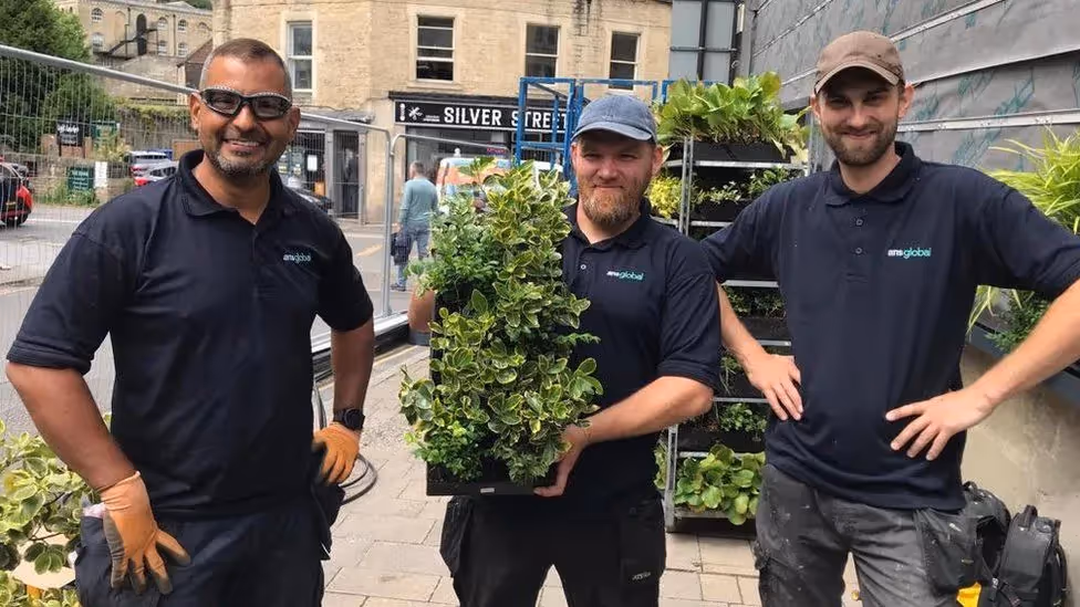 Three landscaping workers with plants on urban street, wearing black shirts