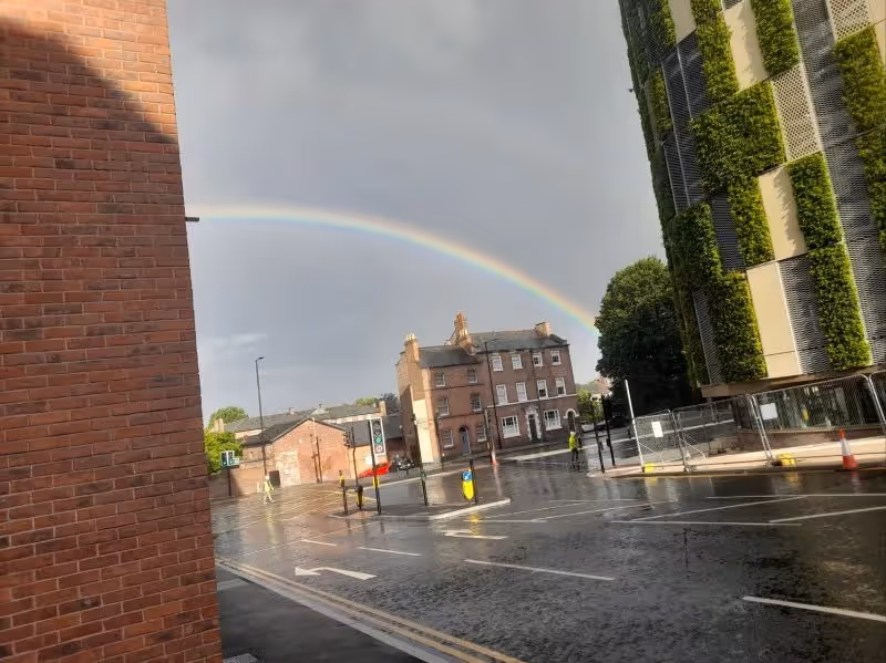 Rainbow arches over wet city street with brick buildings and green wall