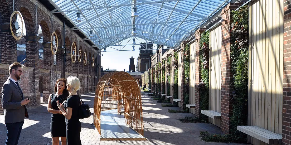Glass-roofed corridor with people, wooden arch, and vertical gardens
