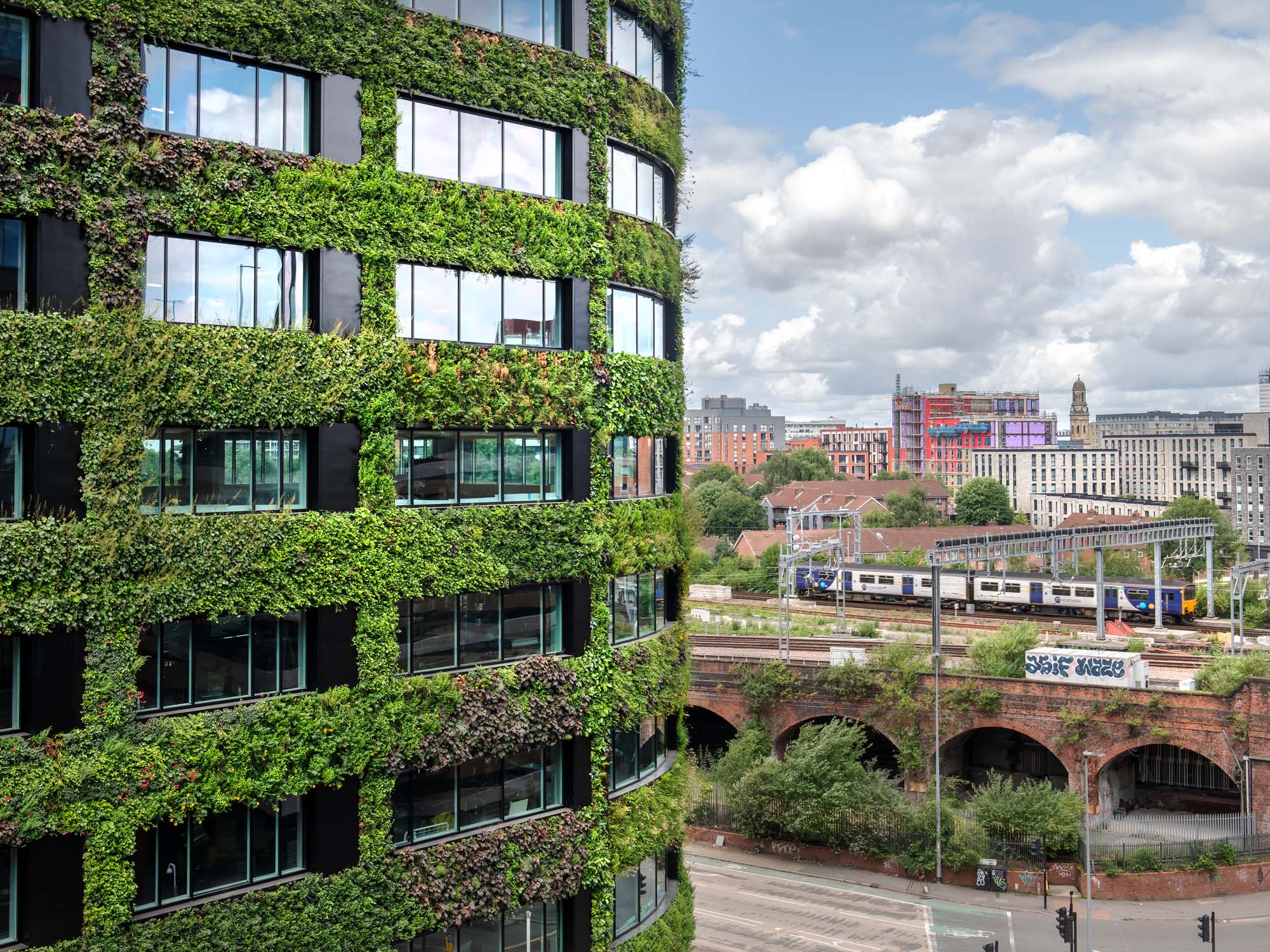 Green-covered modern building near train tracks with urban landscape backdrop
