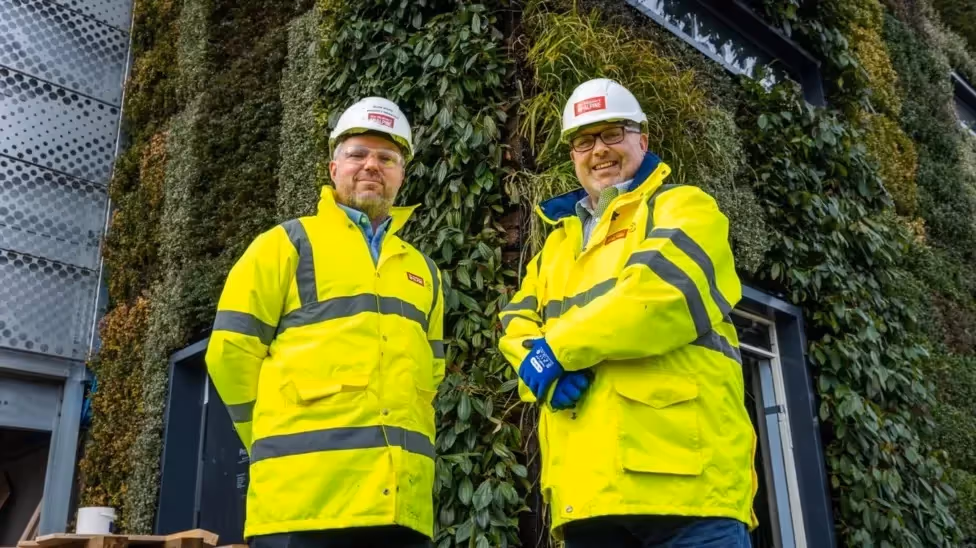 Two construction workers in safety gear stand in front of green wall