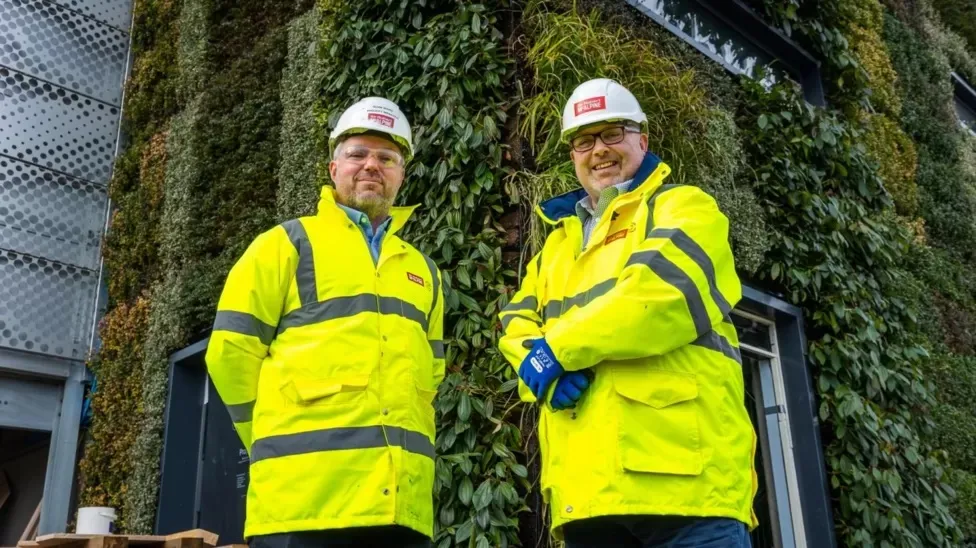 Two construction workers in safety gear stand in front of green wall