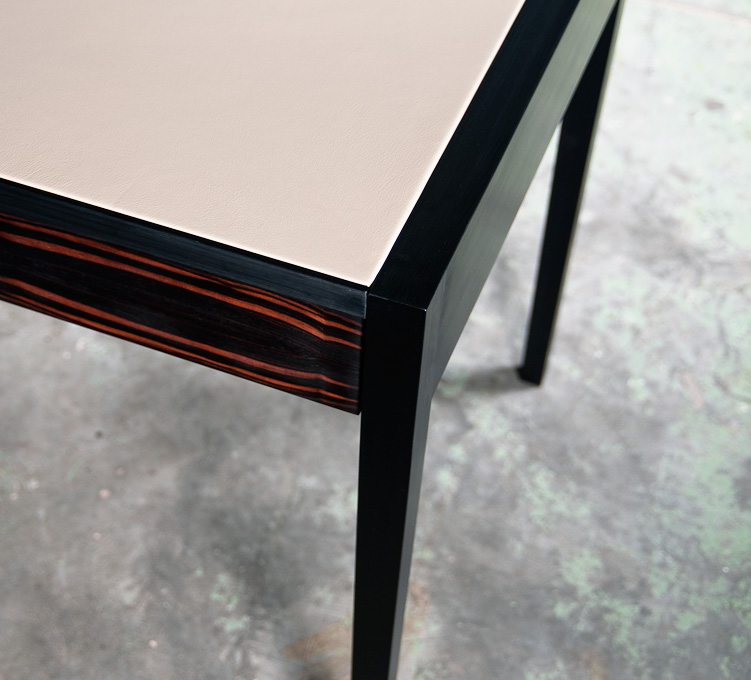 Close-up of a modern table corner with a beige leather top, black frame, and striped wooden apron on a concrete floor.