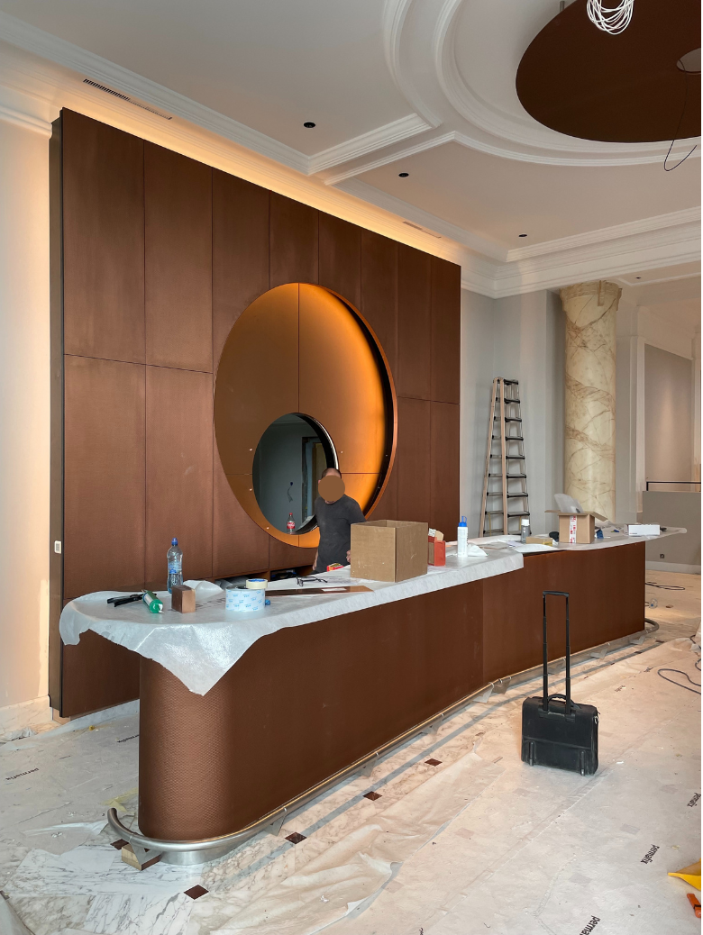 A modern reception desk under construction with brown panels and a large round reflective mirror set into a brown wall, a worker in a black shirt, a ladder, boxes, and construction materials scattered around.