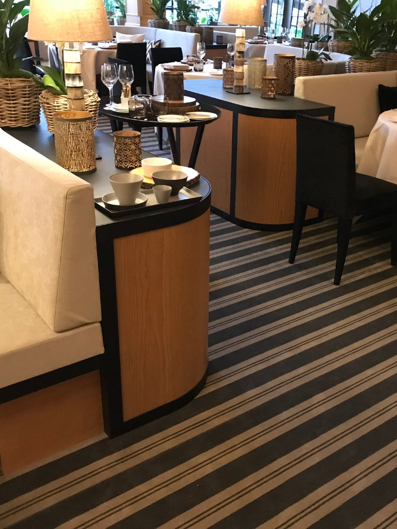 Cozy restaurant interior with wooden and black furniture, tables set with glassware, decorative lamps, and plants on a striped carpet.