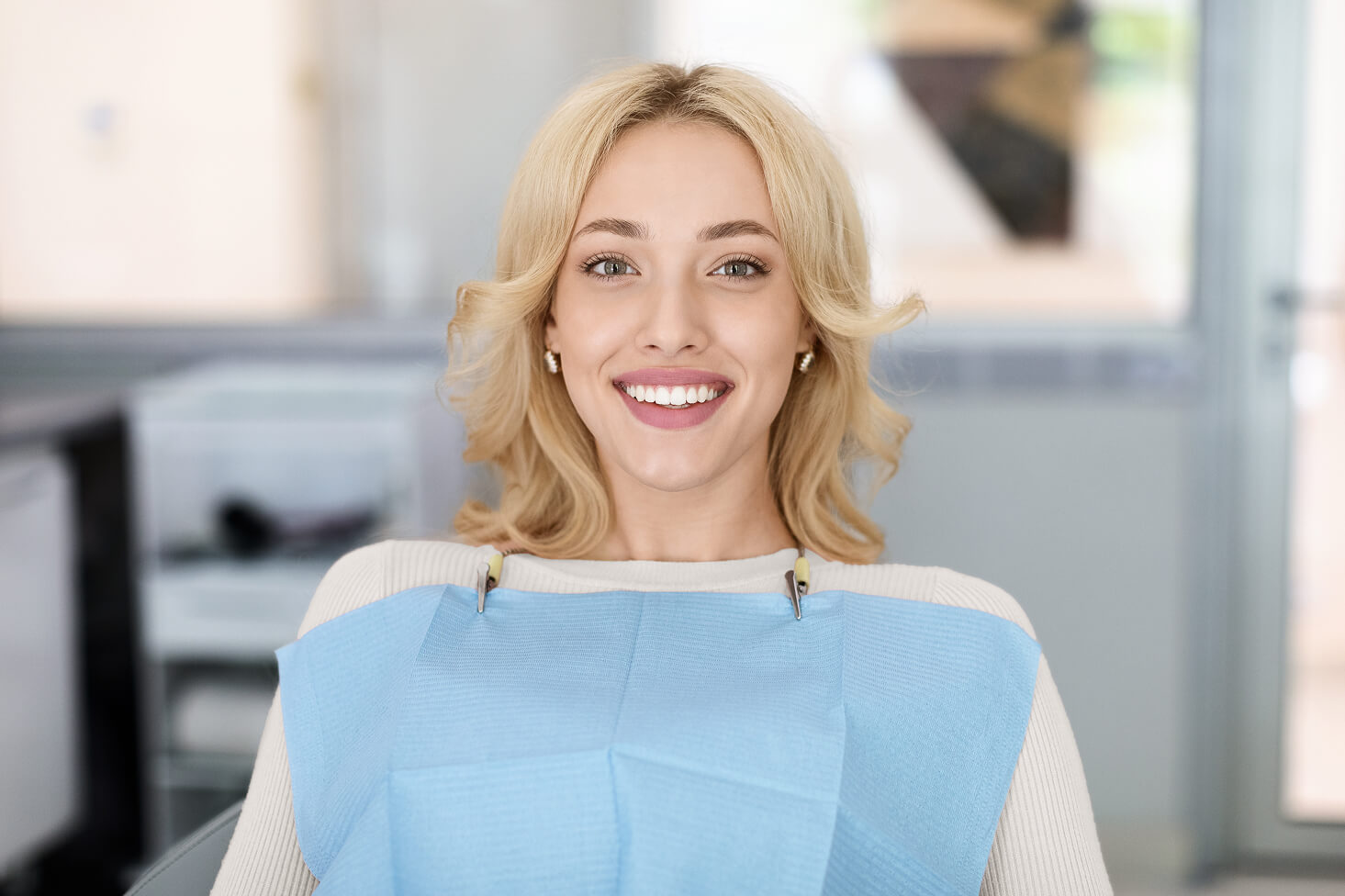 Smiling blonde woman sitting in a dental chair wearing a protective bib, looking relaxed and ready for her appointment at a modern dental office.
