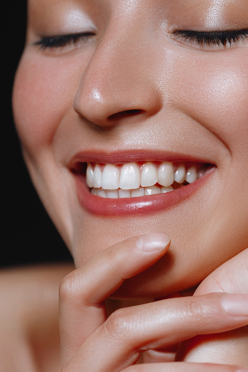 Close-up of a young woman smiling with bright white teeth, eyes closed, and hands gently touching her face, showcasing healthy gums and cosmetic dentistry results.