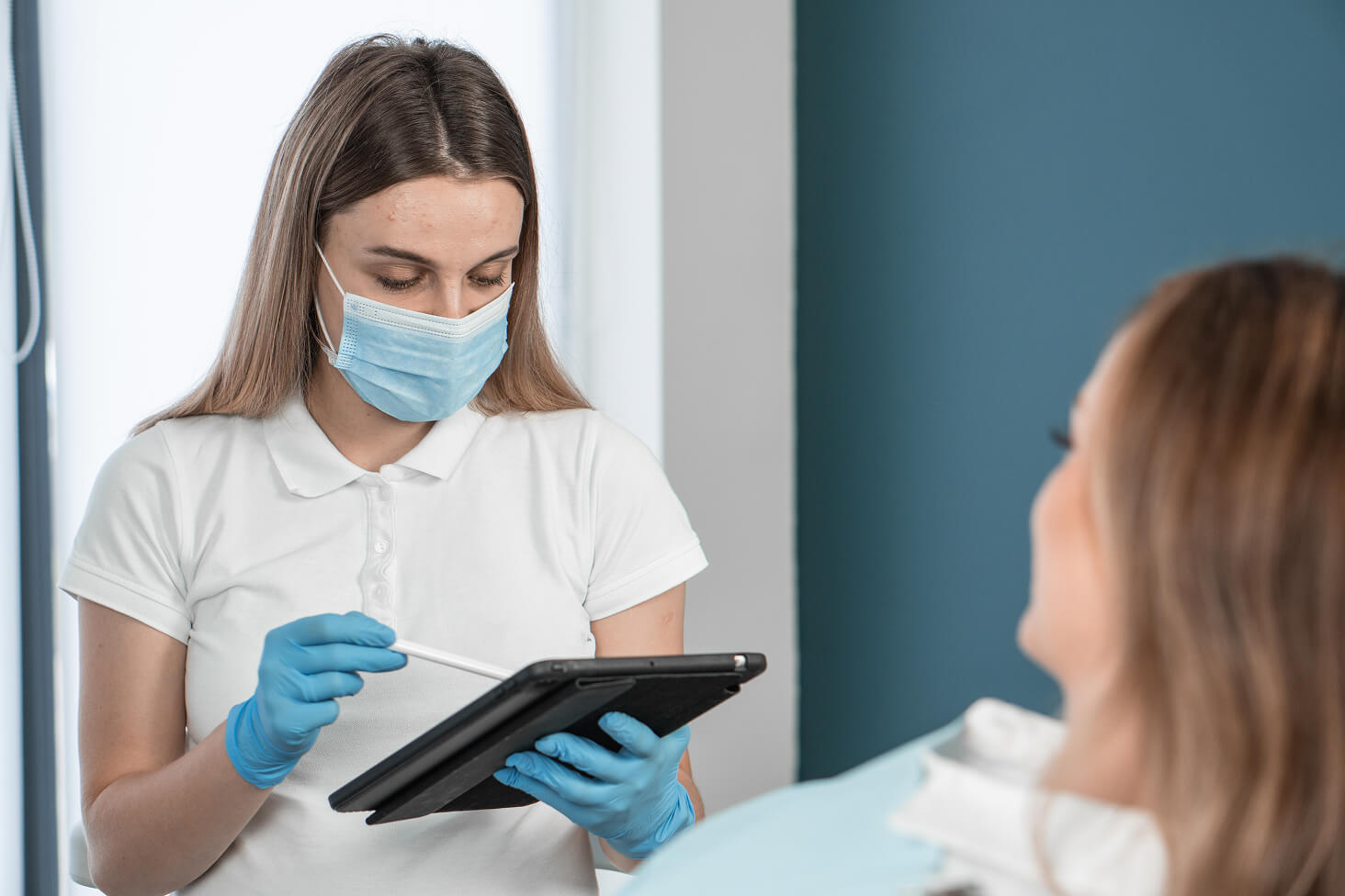 Female dentist wearing mask and gloves holding a digital tablet while discussing treatment options with a seated patient in a dental clinic.