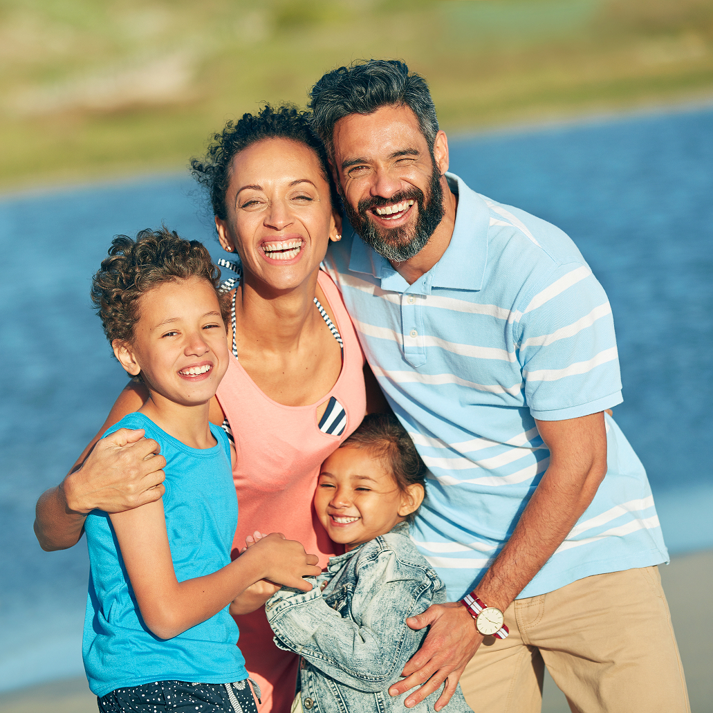 Smiling family of four outdoors with parents hugging their two children, representing healthy, happy smiles.