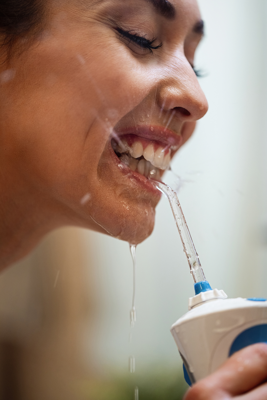 Close-up of smiling woman using a water flosser to clean between her teeth, water dripping around her mouth.