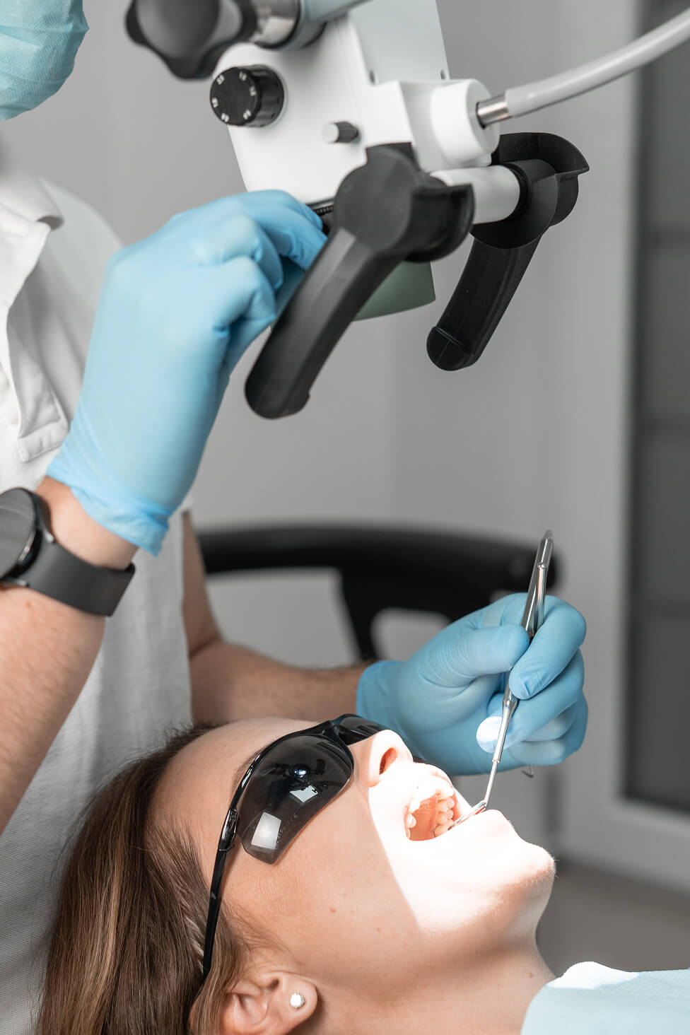 Female dental patient wearing protective glasses receiving a procedure under a dental microscope while the dentist works with precision instruments.