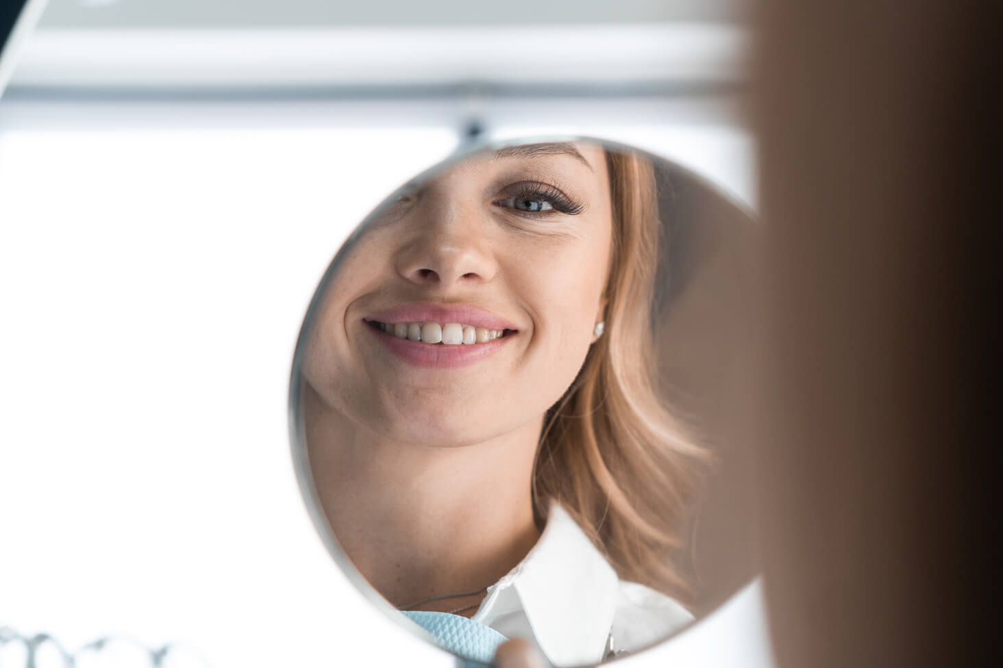 Smiling woman looking at her reflection in a round dental mirror, admiring her healthy teeth after a dental procedure.