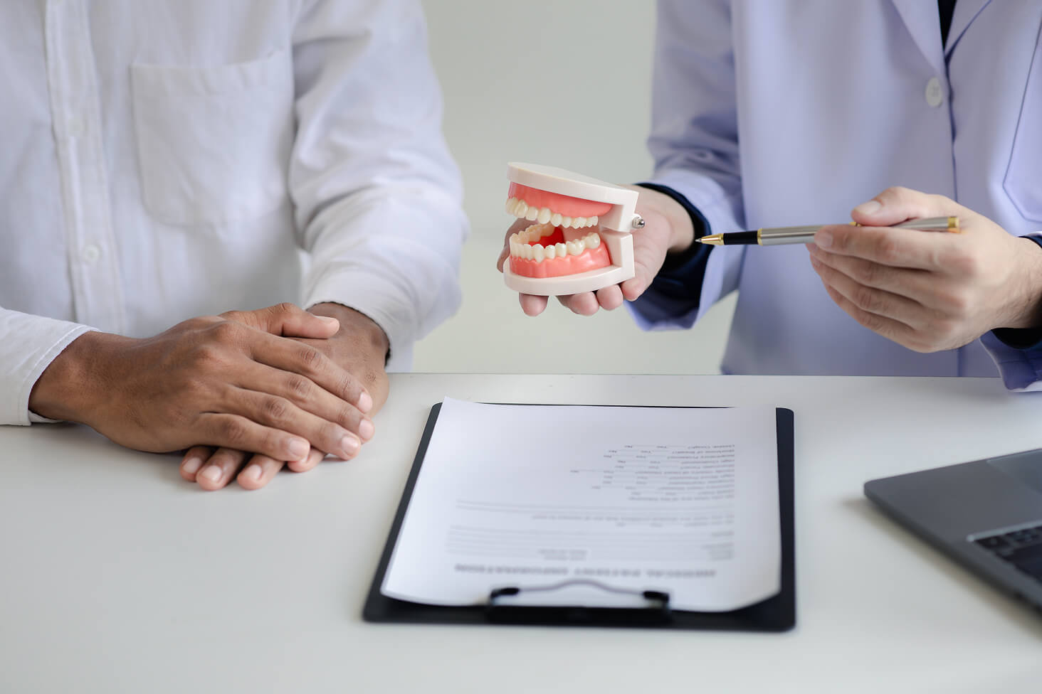 Two dentists in white coats discussing treatment notes with a clipboard and a dental model on the table during a consultation.