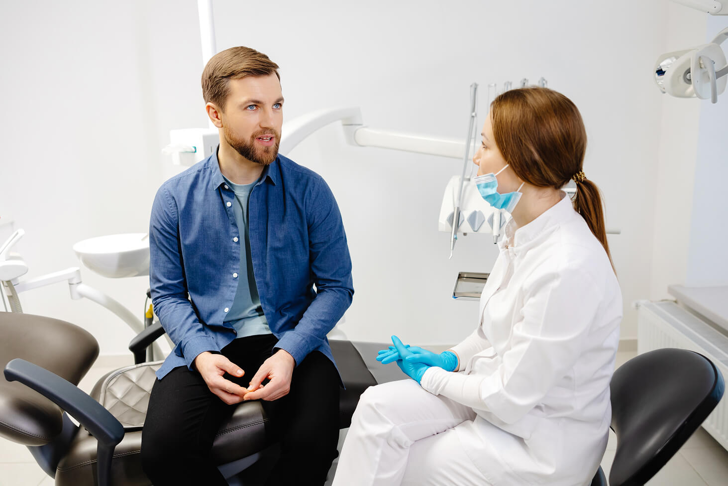 Male patient in blue shirt sitting in a dental chair while a female dentist in white coat and face mask discusses treatment options.
