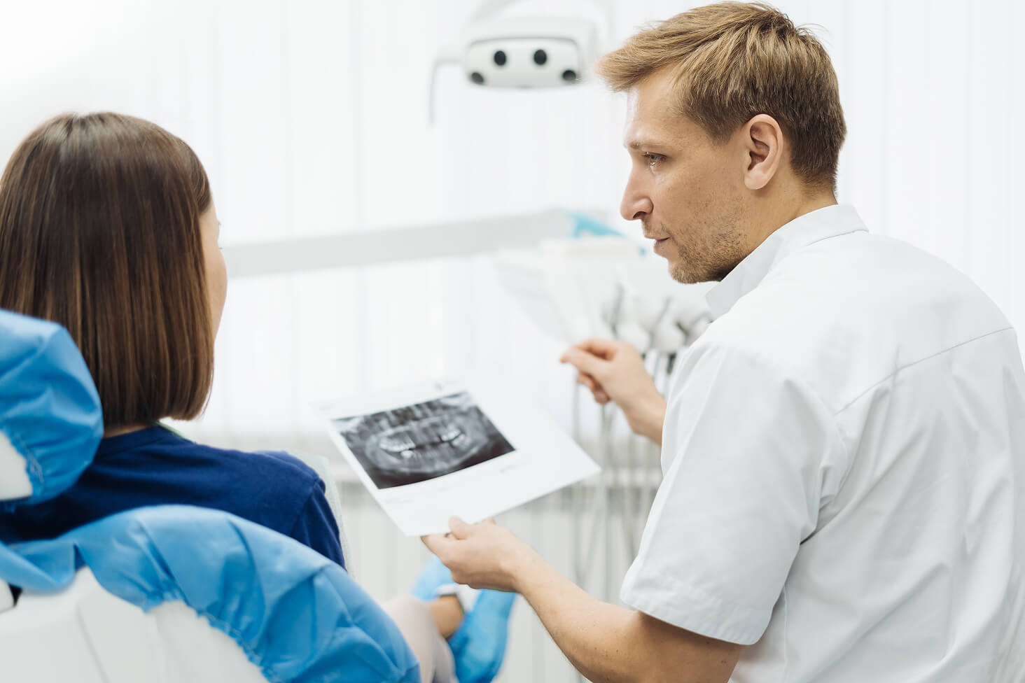 A male dentist in a white coat discussing treatment options with a patient in a modern dental office, pointing toward an X-ray on a tablet.