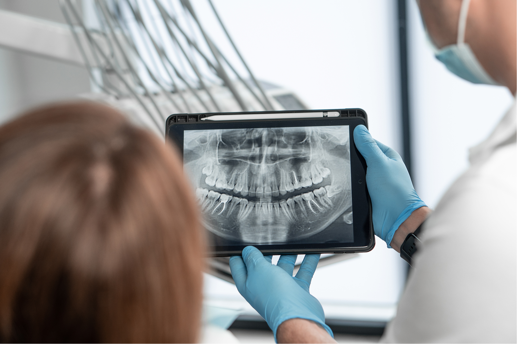 A dentist wearing blue gloves holding up a digital dental X-ray to show a seated patient, explaining the results as part of preventative care.
