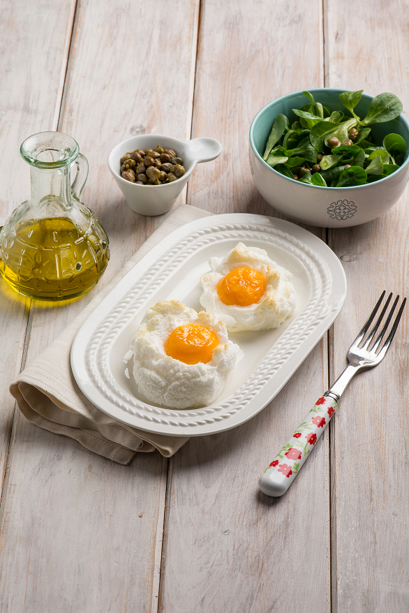 A wooden table setting with a white ceramic plate holding two sunny-side-up eggs, a fork placed on the side, a small bowl of broccoli, a glass bottle of olive oil, and a dish of capers, styled for a clean and healthy breakfast scene.
