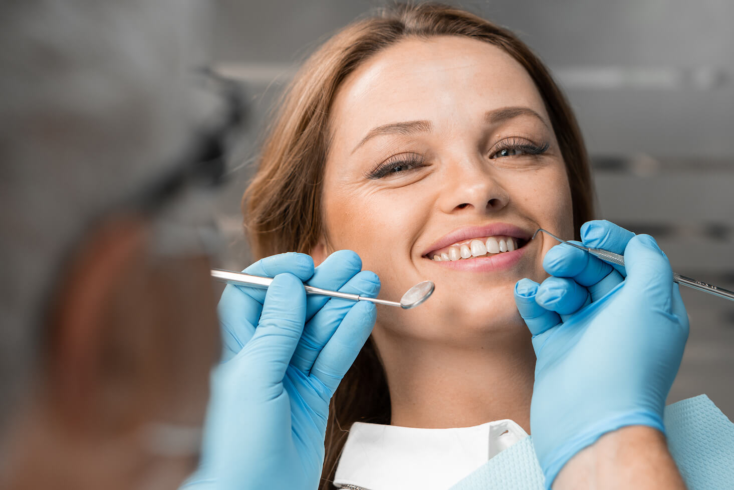 A female dental patient reclines in the dentist’s chair, smiling as a dentist wearing blue gloves uses a dental mirror and scaler to examine her teeth. The patient looks relaxed and comfortable during the oral checkup.