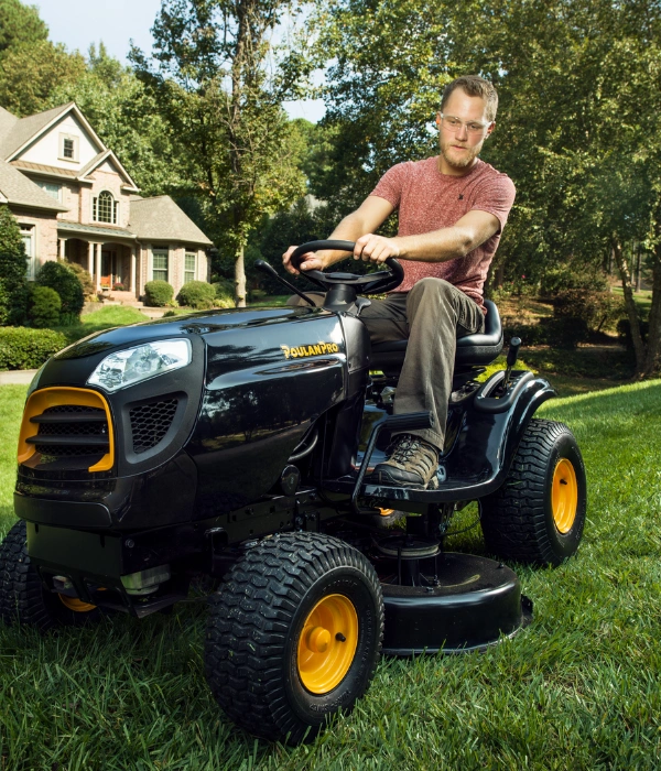 A man wearing safety glasses riding a black Poulan Pro riding lawn mower on a large lawn with trees in the background.