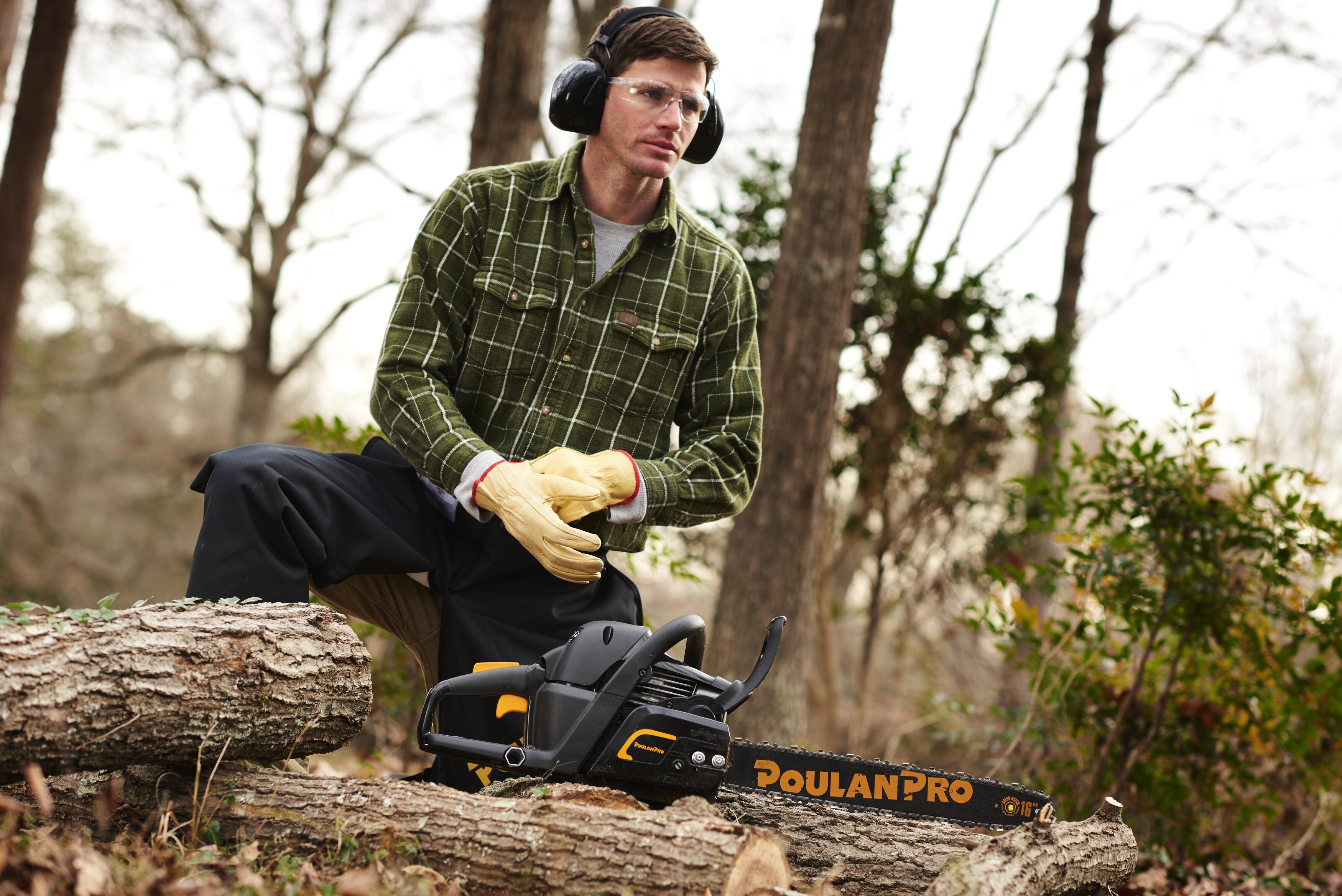 Man in full safety gear operating a Poulan Pro chainsaw outdoors by a log.