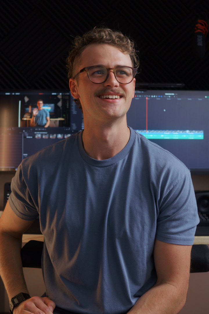 Man in glasses and a blue t-shirt sitting in front of a video editing screen.