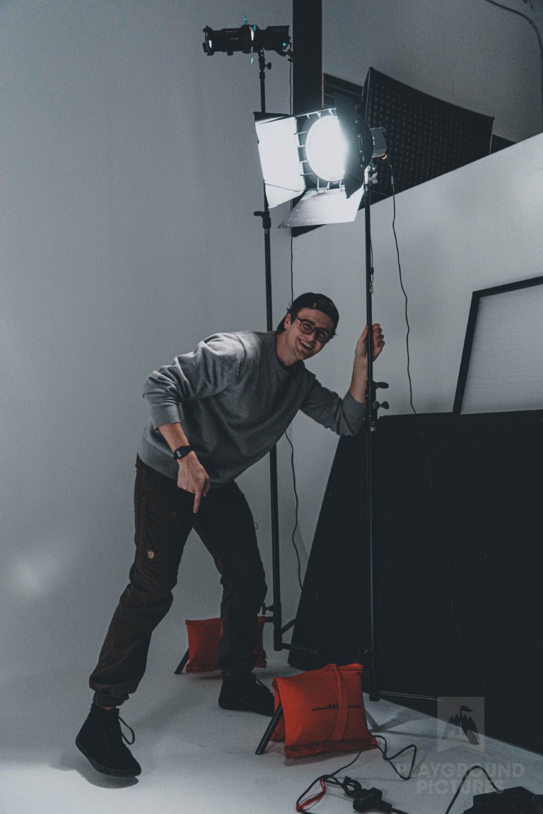 Man in gray sweater and glasses adjusting a studio light on a stand with sandbags on the floor.