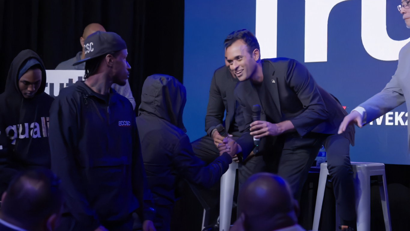 Man in a black suit holding a microphone bending to shake hands with a person wearing a hooded jacket in a dimly lit event.