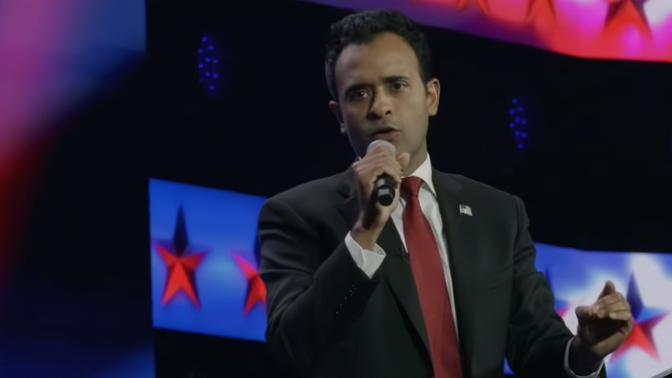 Man in a black suit with a red tie speaking into a microphone on a stage with blue screens displaying red stars in the background.