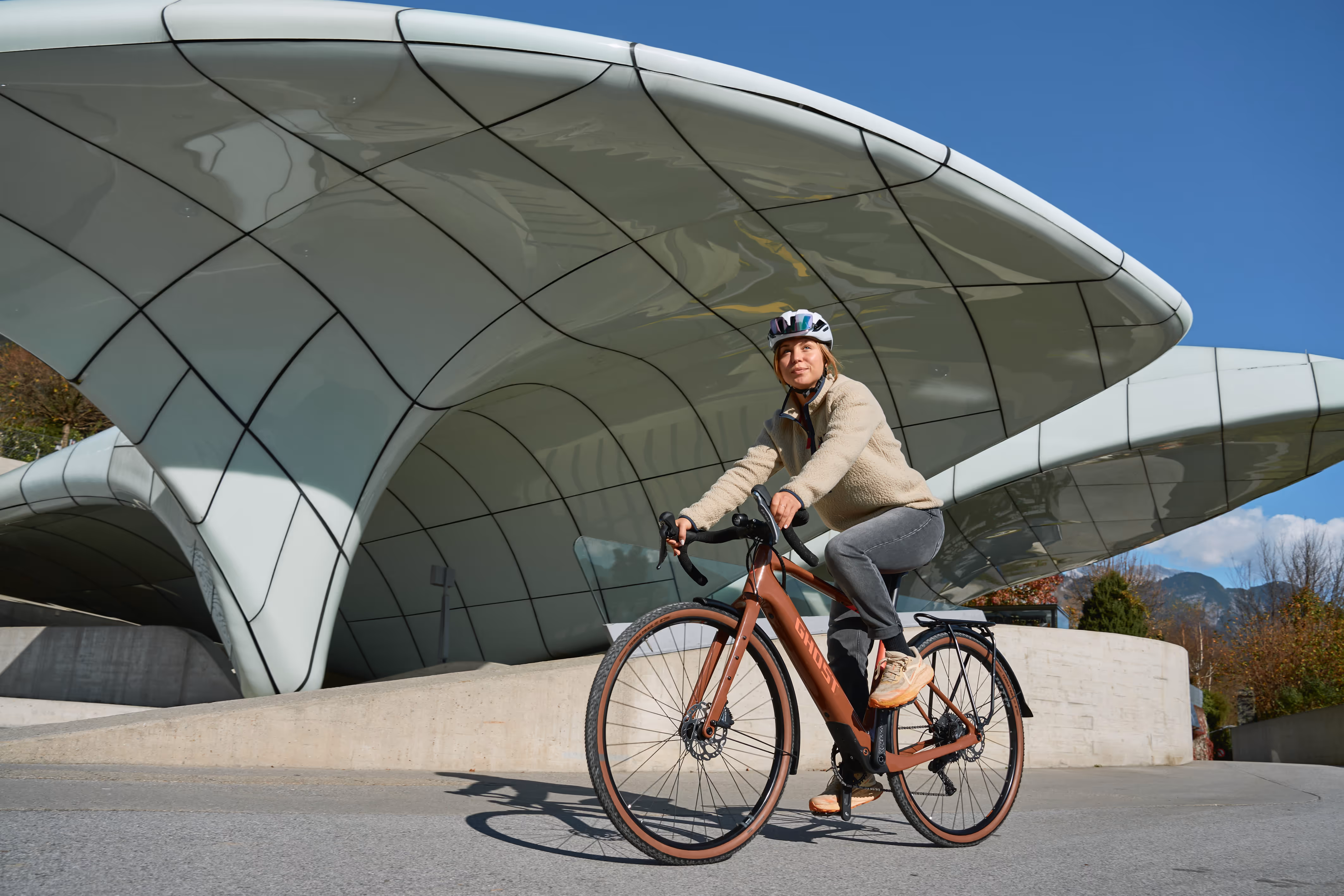 Kobieta na rowerze elektrycznym Ghost przy futurystycznej architekturze
Kobieta w kasku rowerowym na pomarańczowym e-bike'u marki Ghost przy nowoczesnej konstrukcji architektonicznej z zakrzywionymi panelami szklanymi. Widoczne elementy: hamulce tarczowe, rama elektryczna Ghost, beżowy outfit, białe buty. Tło: futurystyczna architektura, błękitne niebo, betonowa nawierzchnia.