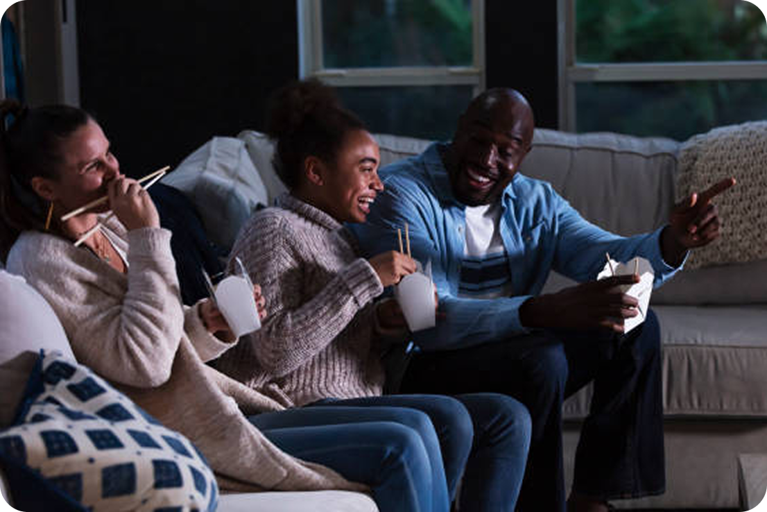 People sitting on the couch eating while looking at the TV and laughing. 