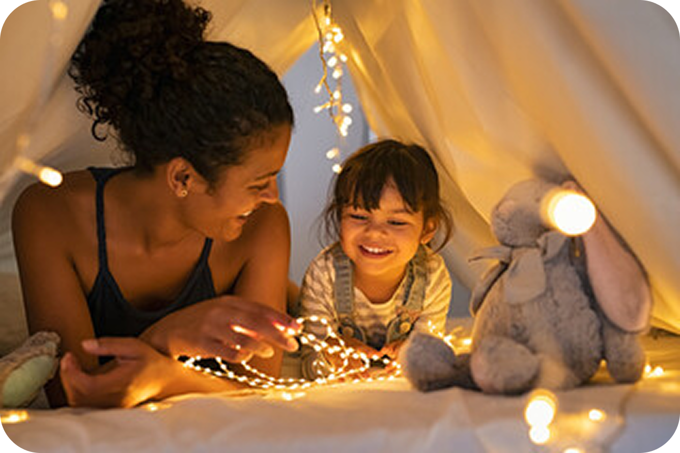Mom and daughter sitting in a tent playing with string lights. 