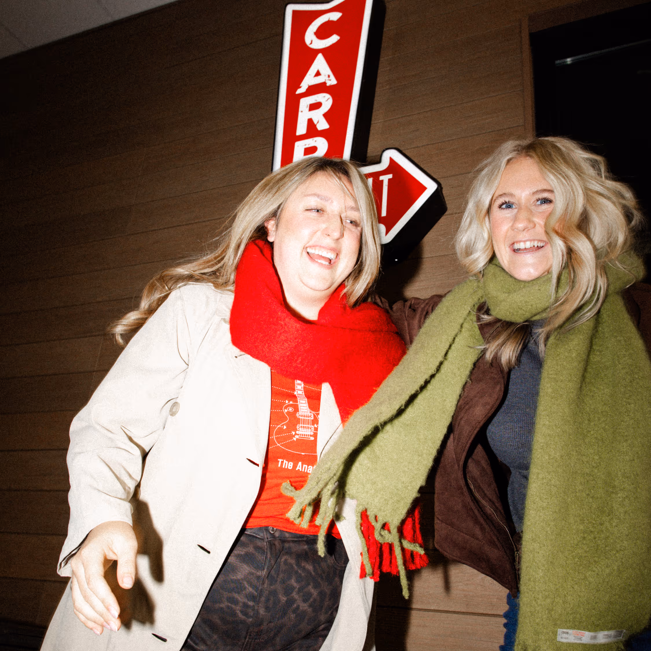 Two smiling women wearing oversized scarves, standing close together in front of a wooden wall with a red vertical sign that reads 'CARD'.