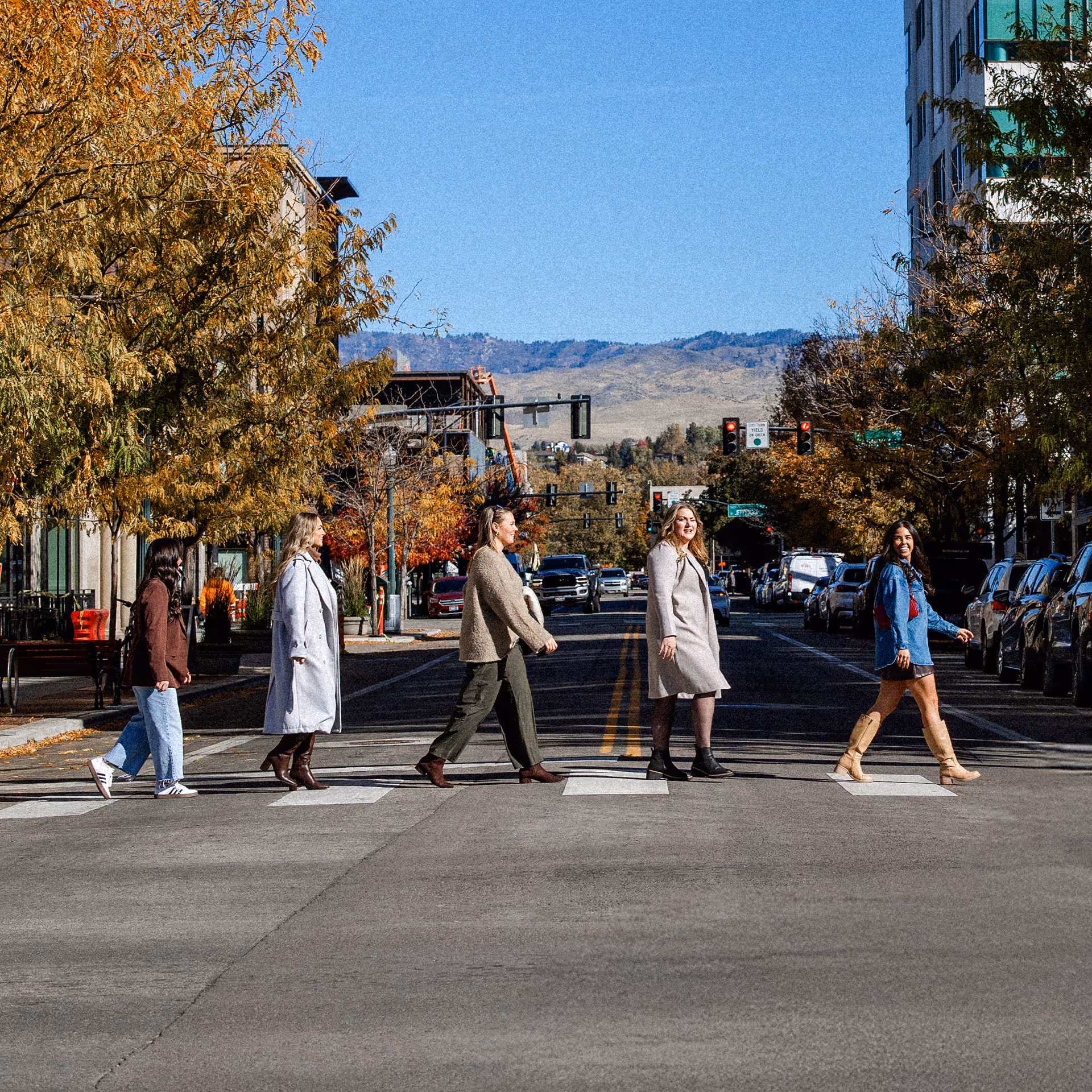 Five people crossing a city street on a sunny day with fall foliage and mountains in the background.