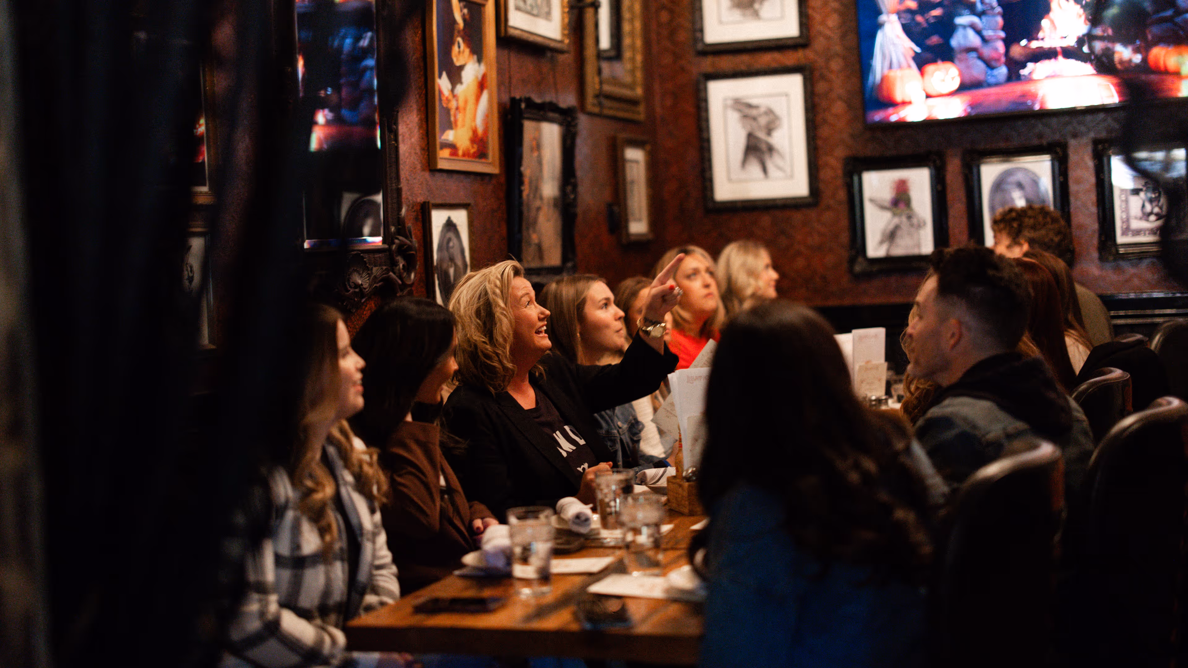 Group of people seated at a wooden table in a warmly lit restaurant with framed artwork on walls, one woman pointing and smiling.