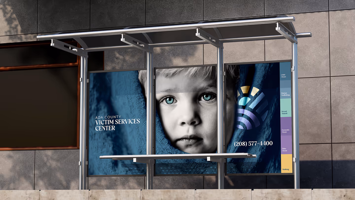 Bus stop ad for Ada County Victim Services Center. Features a somber, monochrome close-up of a child's face, contact number, and colorful vertical info bars.