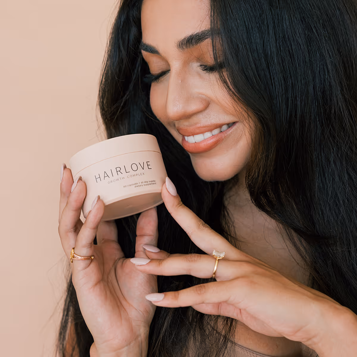 Smiling woman with long, dark hair holds a round container labeled "HAIRLOVE Growth Complex." She wears gold rings against a soft peach background.
