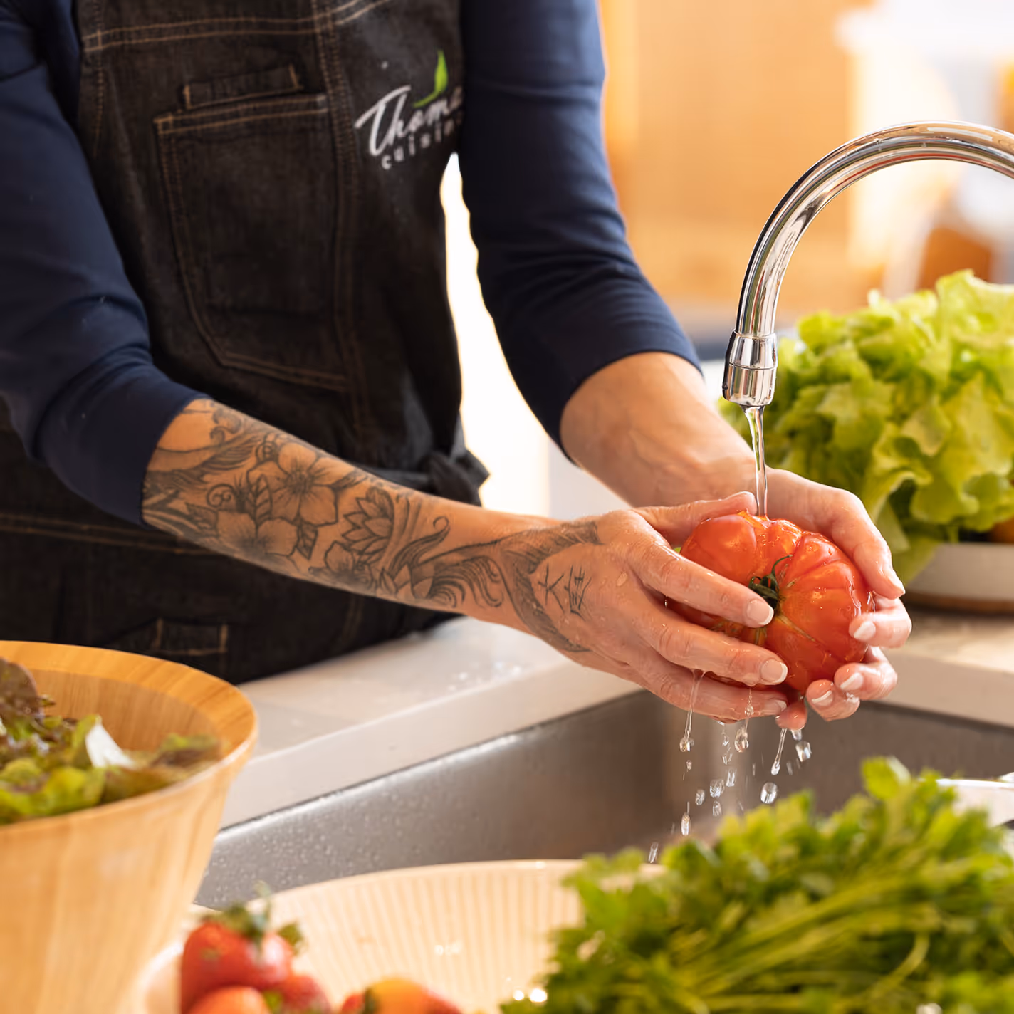 A person with tattooed arms wearing an apron washes a large tomato under a kitchen faucet, surrounded by bowls of fresh lettuce and vegetables; a calm, focused atmosphere.