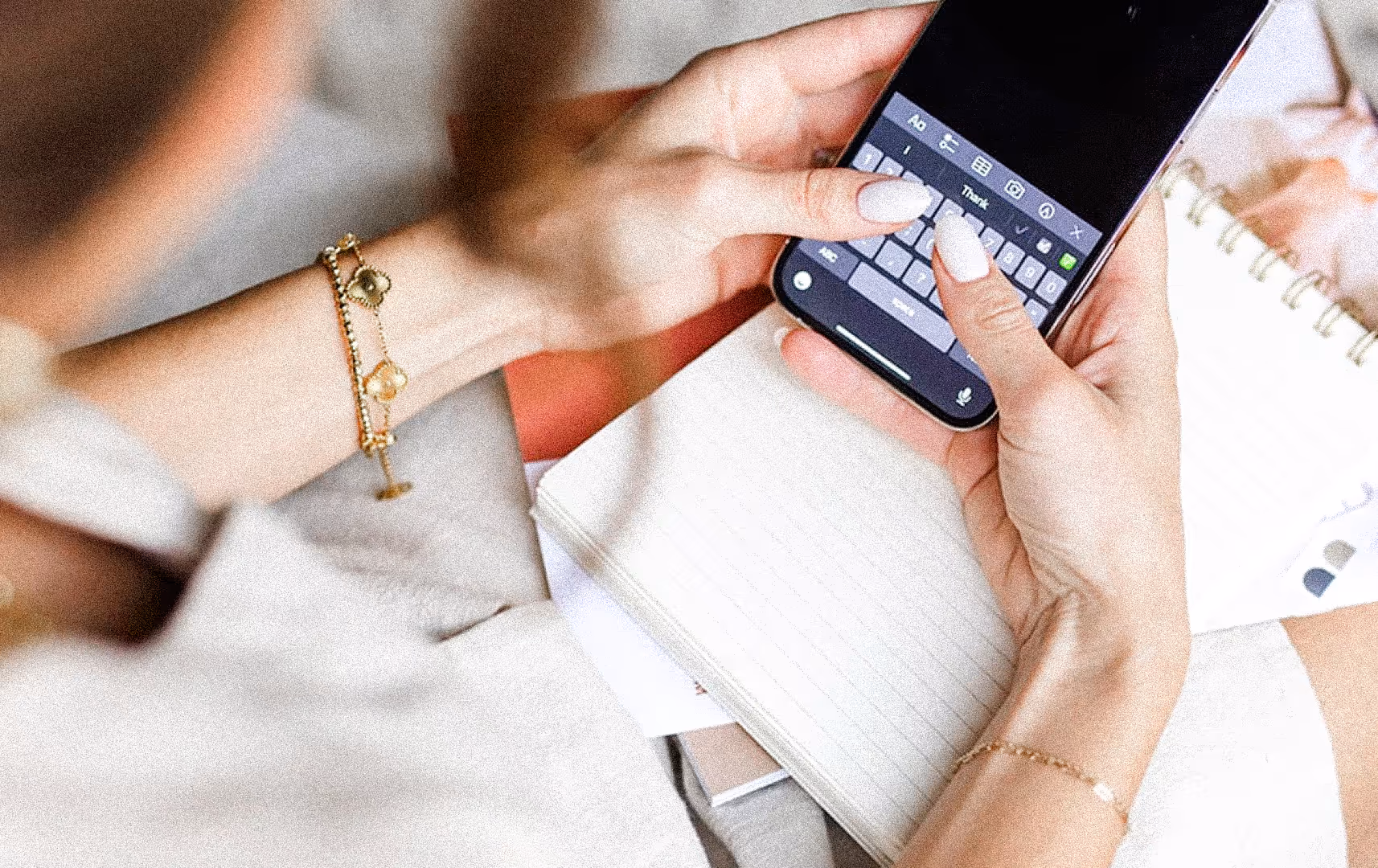 A woman types on a smartphone over a lined notebook. Her gold bracelets are visible. The scene conveys focus and multitasking in a relaxed setting.